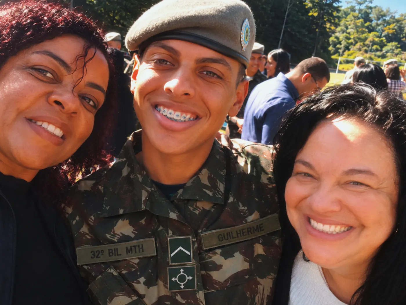 Guilherme Carvalho de Andrade de uniforme e boina do Exército Brasileiro, sorrindo ao lado da madrasta, à esquerda, e da mãe, à direita, durante uma cerimônia militar ao ar livre.